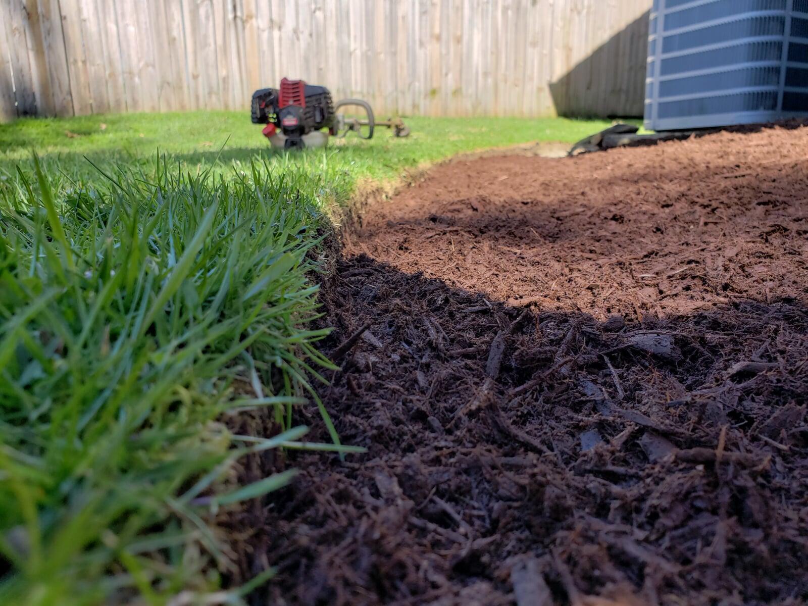A close-up view showcasing a freshly edged landscape bed, highlighting a clean, precise separation between the lush green grass and newly laid dark mulch. A piece of landscaping equipment rests in the background, emphasizing professional maintenance work.