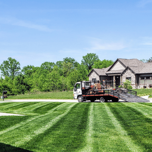 A freshly mowed lawn with clean, distinct stripes, showcasing TruScape's professional landscaping service. A landscaping truck with equipment is parked on the street in front of a residential property, highlighting a well-maintained lawn on a sunny day.