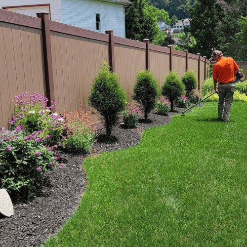 A landscaping professional wearing a high-visibility orange shirt carefully trims the edge of a freshly maintained garden bed alongside a neatly mulched flowerbed. The garden features vibrant flowers and neatly shaped shrubs along a wooden privacy fence, highlighting meticulous attention to detail and quality workmanship in a residential setting.